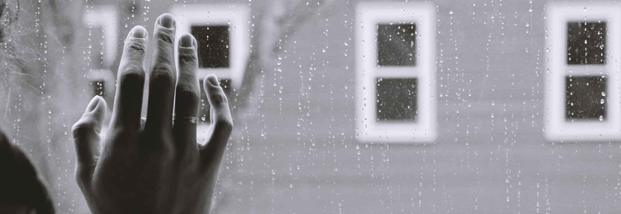 someone’s hand placed on a rainy window, looking out at other windows